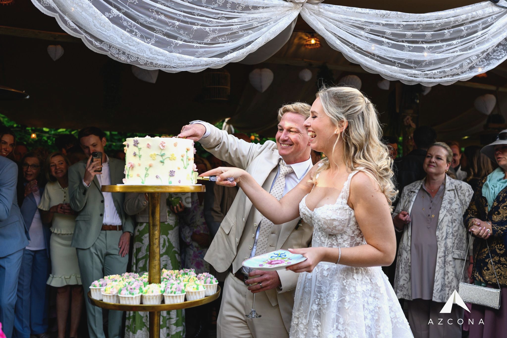 A newly married couple cutting their Butter Up floral wedding cake above a display of matching cupcakes